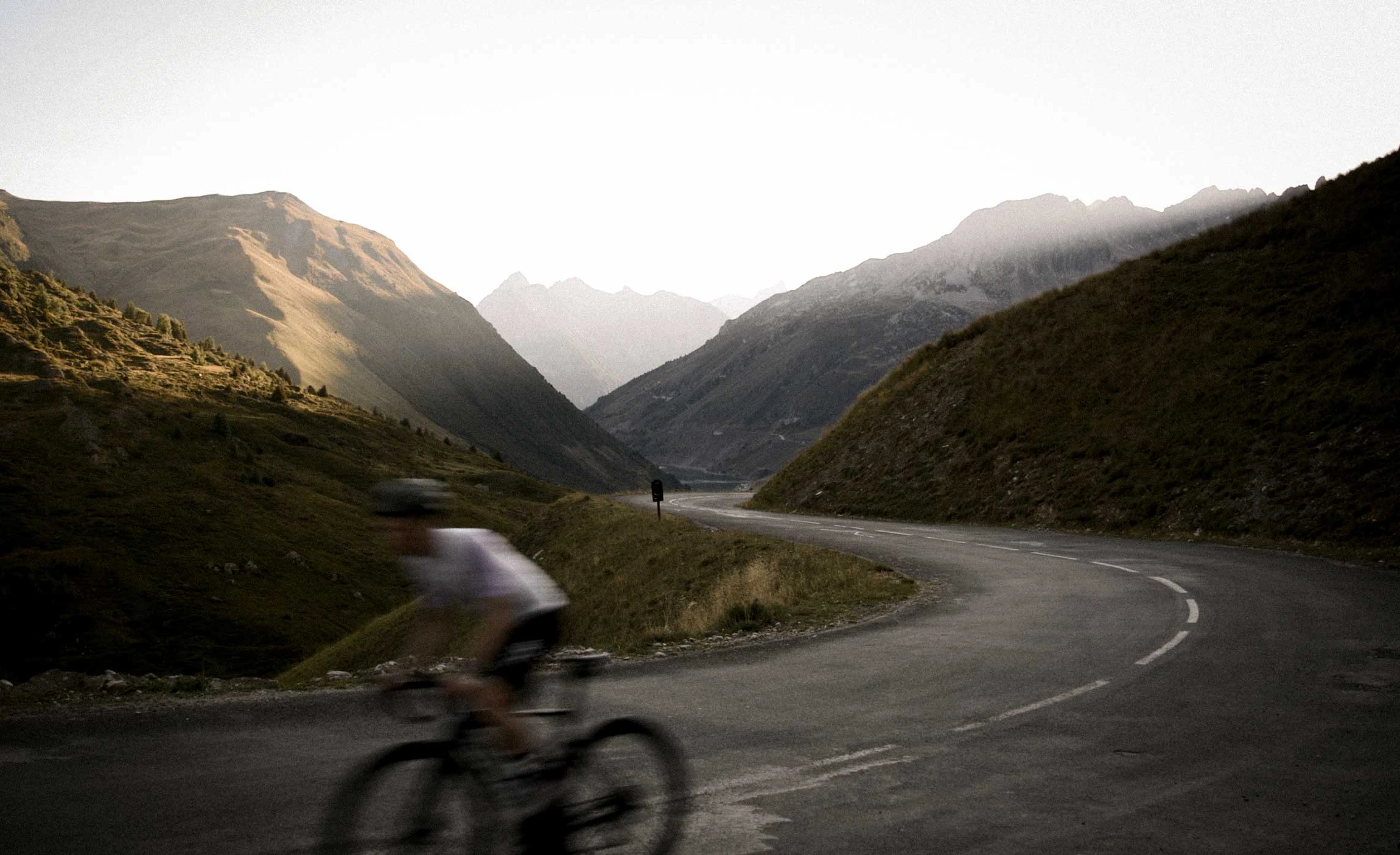 Cyclist riding a winding mountain-pass road through a misty alpine valley at dawn